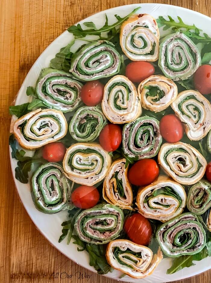 Tortilla pinwheel recipes show Appetizer pinwheels on white plate with grape tomatoes and a bed of arugula. Bamboo cutting board in the background. 