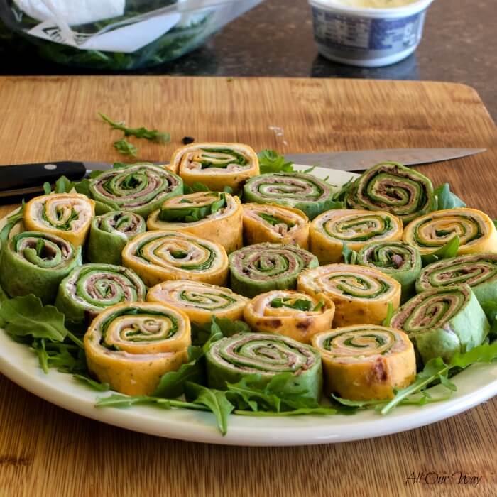 Tortilla pinwheel recipes show spirals on a bed of arugula on a white plate set on a cutting board. A carving knife is in the background. 