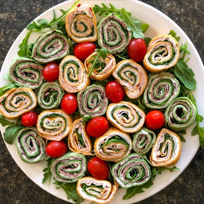 Red and Green Tortilla pinwheels on a white platter with red cherry tomatoes and a sprinkling of green arugula around the platter.