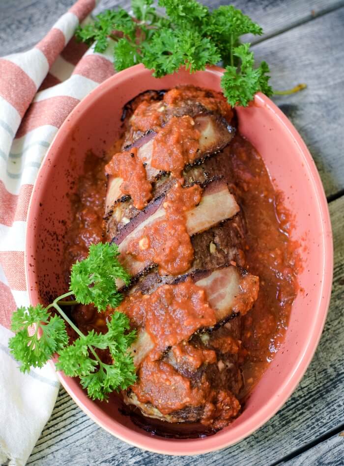 Polpettone Italian stuffed meatloaf in casserole dish with strips of bacon and spoonfuls of marinara sauce on top. Sprigs of green parsley next to meatloaf.