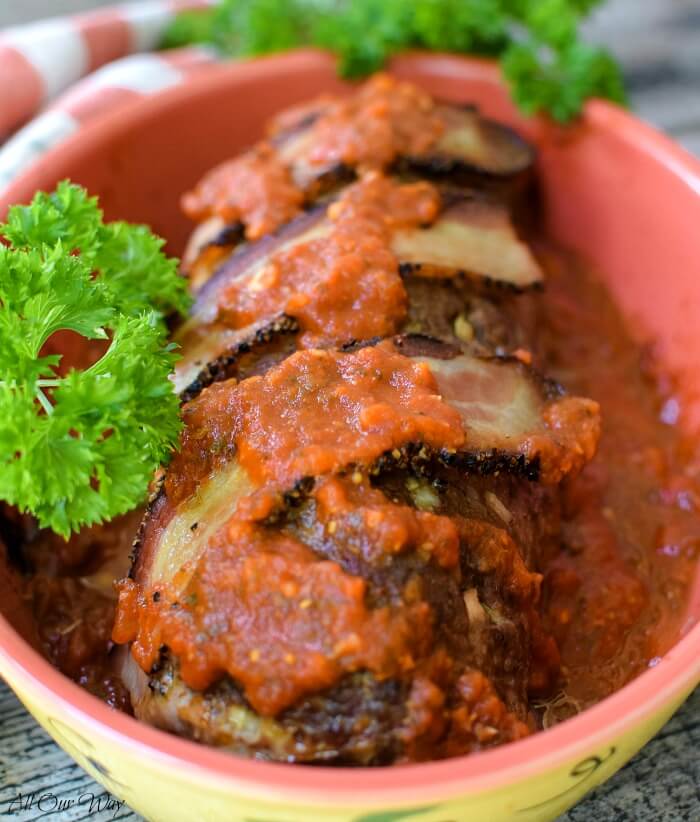 Polpettone Italian stuffed meatloaf is a red casserole dish with green parsley on the side. Bacon and marinara sauce tops the meatloaf. 