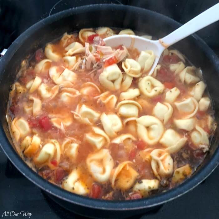 Large pot of Chicken Spinach Tortellini Soup simmering. Large white spoon in pot stirring in the cheese tortellini.