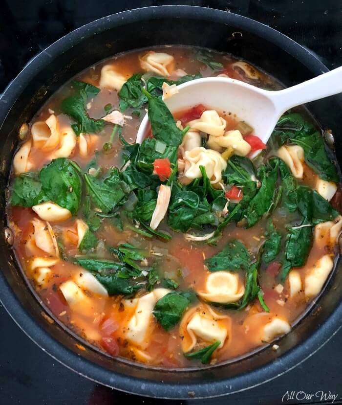 Chicken Spinach Tortellini Soup in large black pot with white spoon stirring in the fresh baby spinach. 