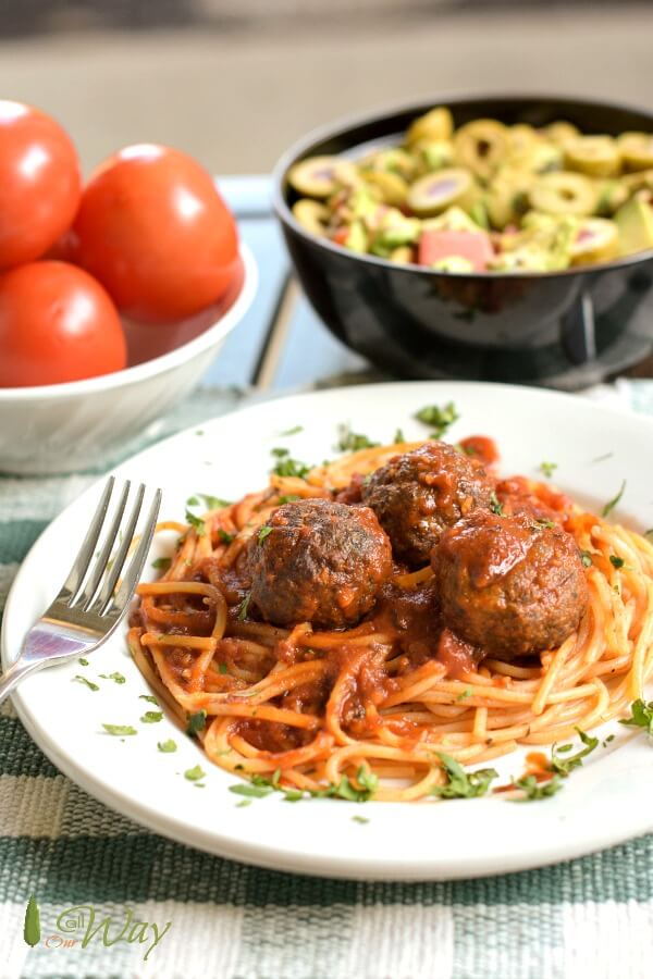 A table set with a white plate with three baked venison meatballs with spaghetti covered in a red sauce. Chopped basil is sprinkled on top. Fork is on left side, a white bowl of red roma tomatoes are behind the plate and next to it is a brown pottery bowl with a tossed salad. 