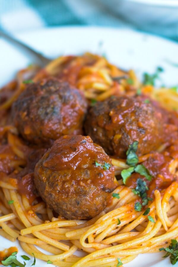 Closeup of three baked venison meatballs on top of spaghetti and covered with a red sauce all on a white porcelain plate and basil leaves sprinkled on top. 