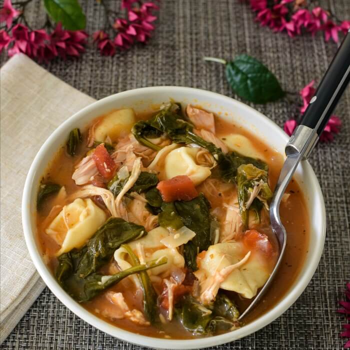 White bowl full of chicken spinach tortellini soup on woven brown mat with ecru linen napkin and purple flower buds on mat. Black-handled soup spoon in the bowl.