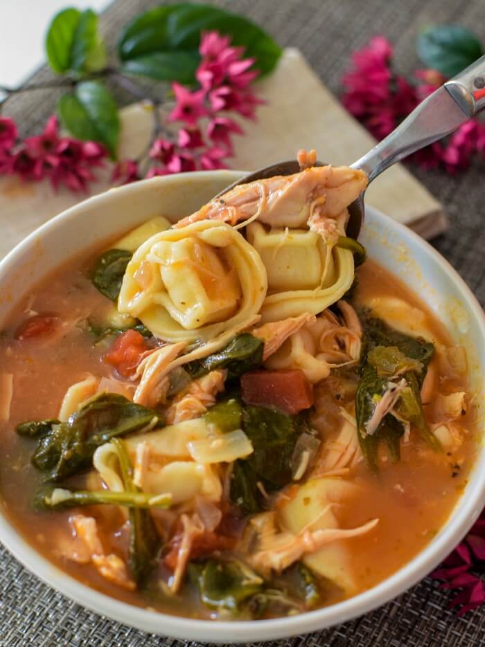 Bowl of chicken spinach tortellini soup with a spoonful of tortellini. Purple flowers in background. 