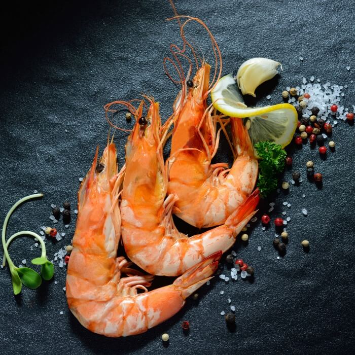 Three large cooked shrimp on black slate stone with sliced lime and peppers strewn around.