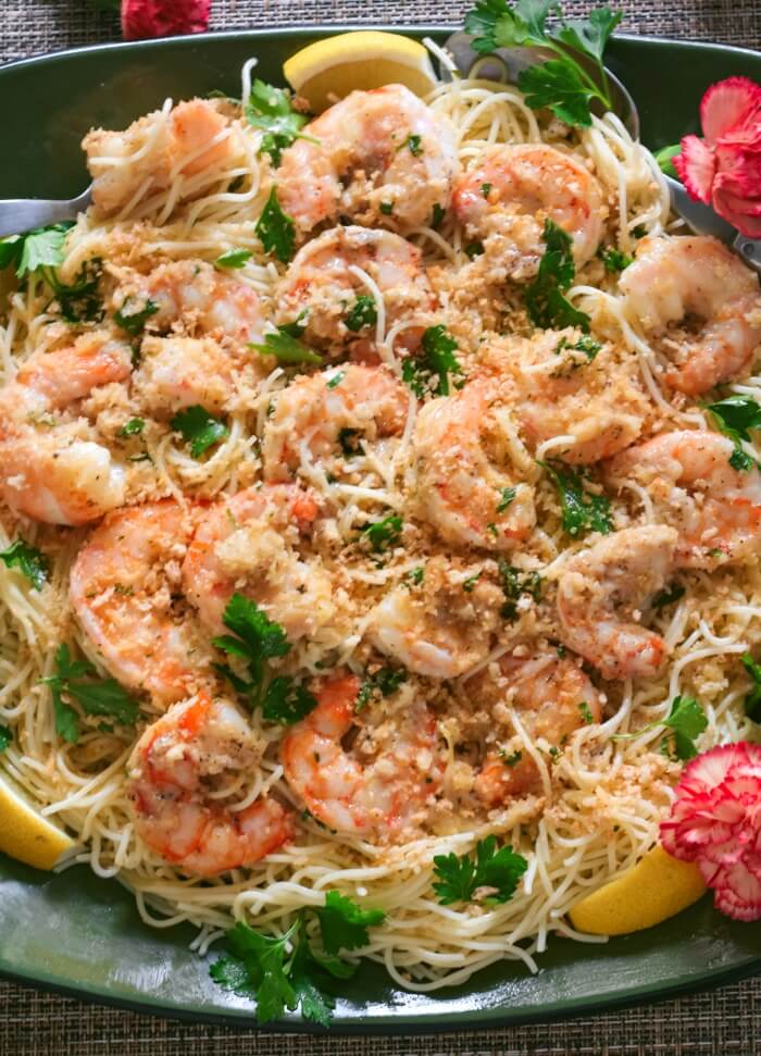 Easy shrimp scampi on a bed of angel hair pasta served on a platter with parsley and lemon with pink flowers in scene.