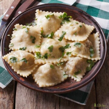 Homemade Italian ravioli with meat and cheese filling sprinkled with green parsley in a brown rustic bowl with wood handled fork and knife on a green and white plaid napkin on top of rustic wood table.