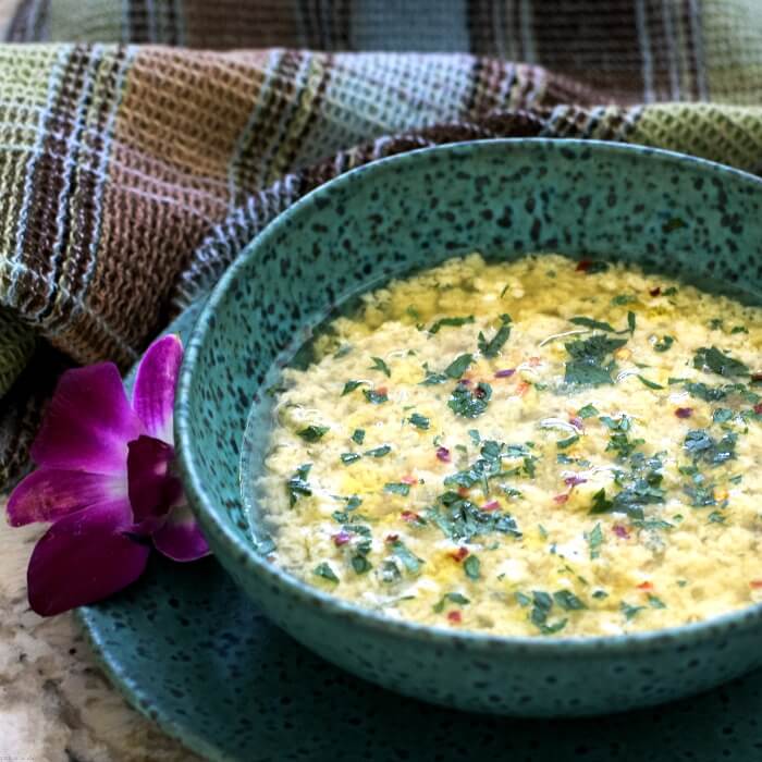 Stracciatella all Romana is a broth soup in a turquoise speckled bowl on matching plate with purple orchid on the left side and a brown, blue, green tea towel behind it. 