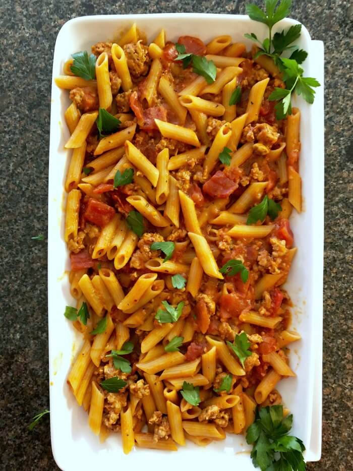 A large white rectangular plate filled with Italian sausage tomato pasta sprinkled with green flat leaf parsley. White platter is on a black and tan speckled marble.