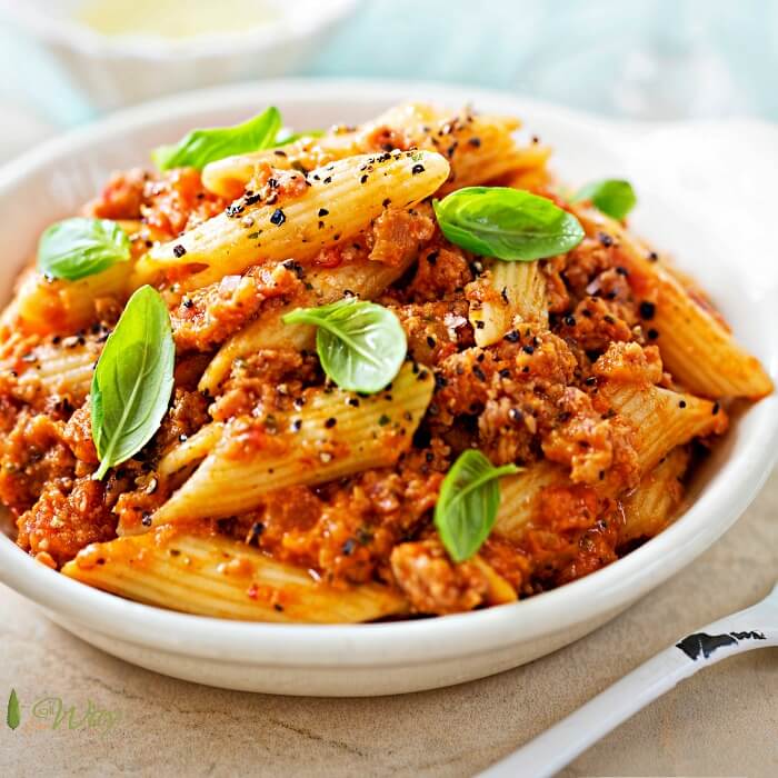 A closeup of Italian sausage penne pasta in a white bowl with a sprinkle of fresh peppercorns and green basil leaves. 