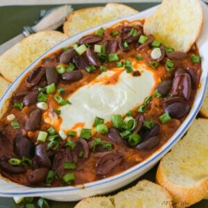 Baked goat cheese dip surrounded by red marinara sauce in a white gratin dish that sits on a dark green platter surround with golden round bread toasted as dippers for the appetizer.