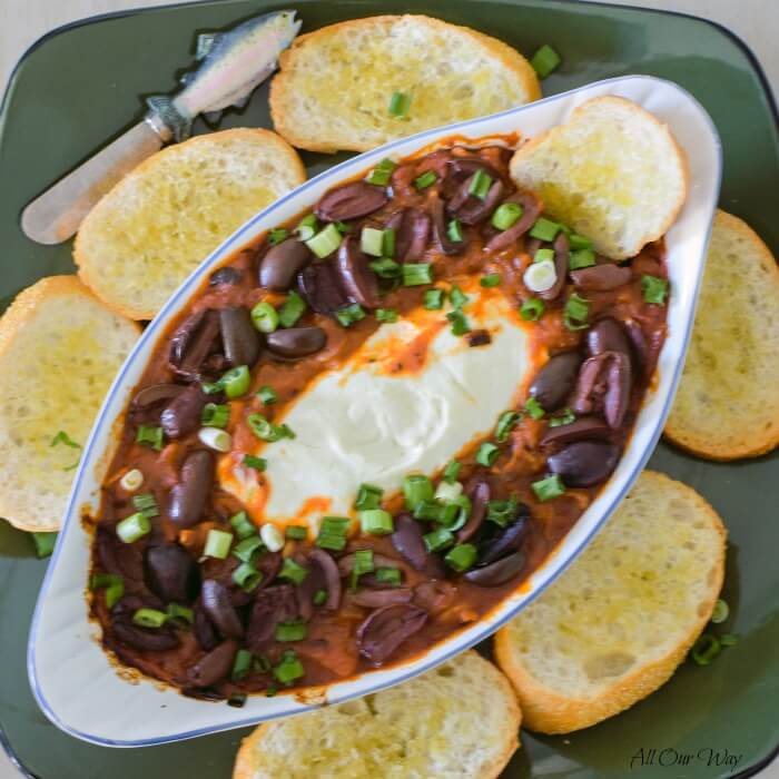 Goat cheese log in the center of a white gratin dish surrounded by marinara sauce with purple kalamata olives and green onion rings sprinkled on top of the red sauce. The gratin dish sits on a green platter with golden toast surrounding the dish and a small cheese knife on the edge. 