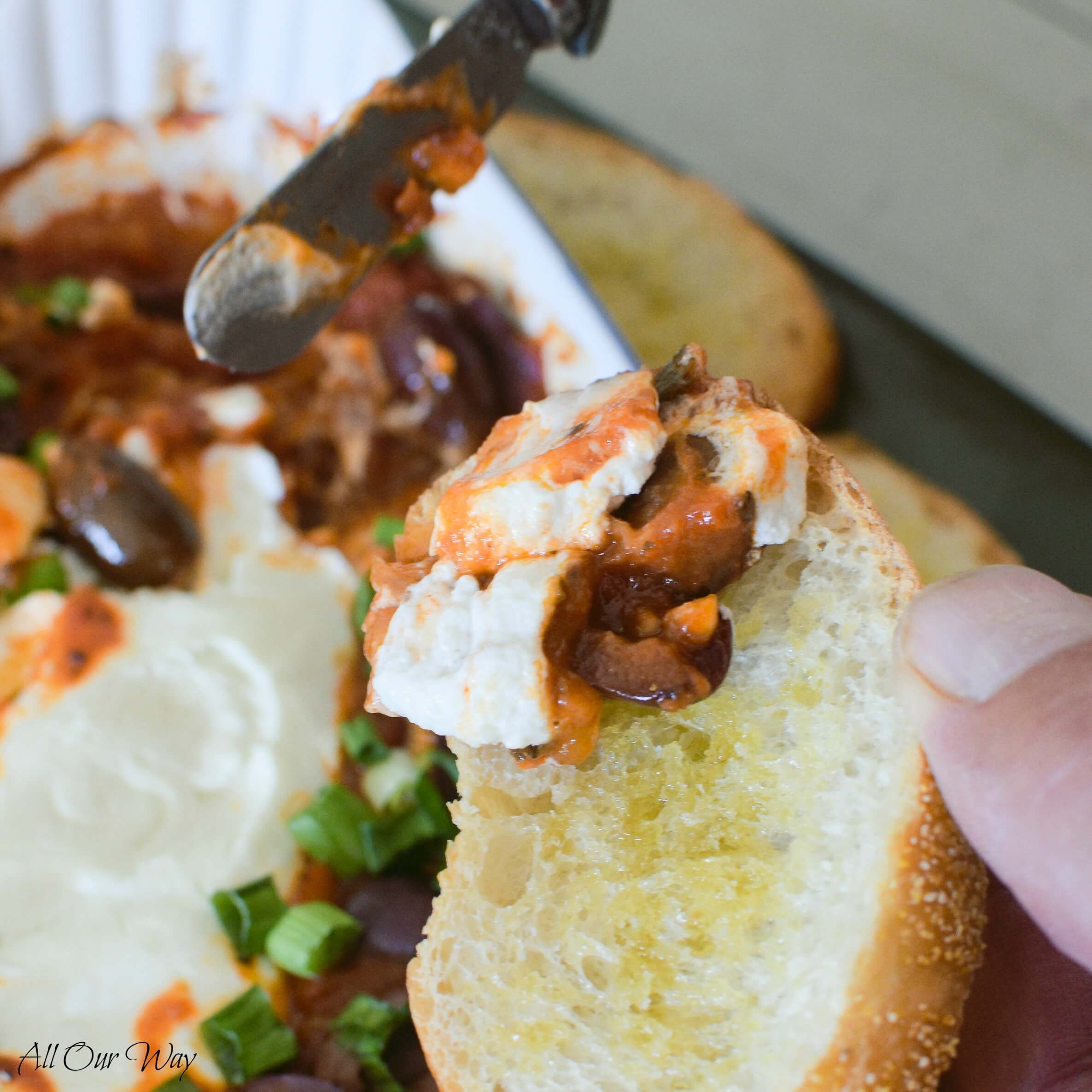 A close-up of man holding toasted bread with a cheese knife putting a dollop of smooth cream cheese and red marinara sauce. White gratin dish with the baked goat cheese marinara dip in the background. 