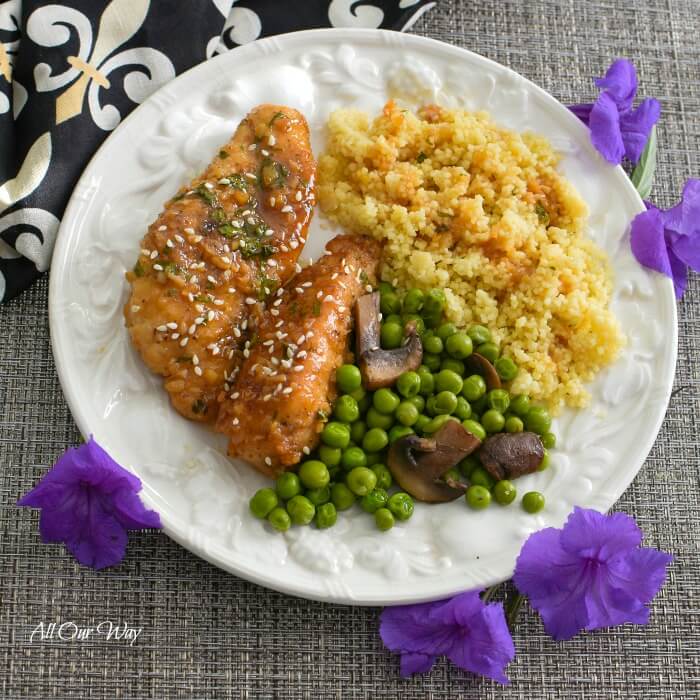 A white plate holds two pieces of honey garlic chicken , green peas with mushrooms, and golden quinoa with honey sauce drizzled on top. Surrounding the plate are 5 purple blossoms and at the upper left hand side is a black napkin with white and gold fleur de lis. 