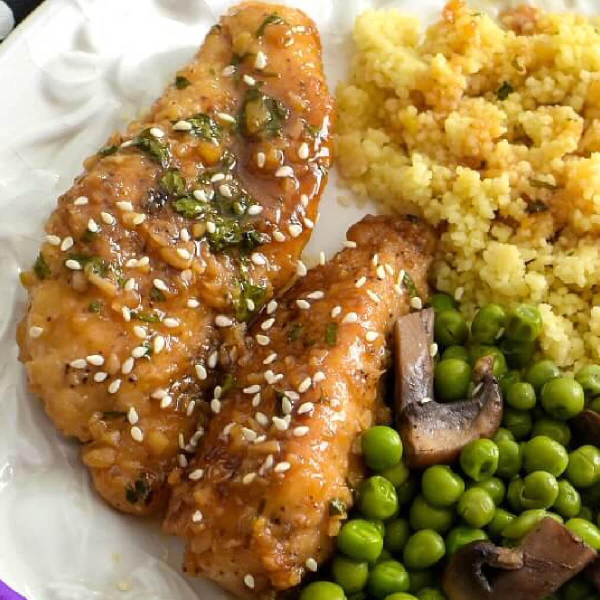 A white plate holds two pieces of honey garlic chicken , green peas with mushrooms, and golden quinoa with honey sauce drizzled on top. Surrounding the plate are 5 purple blossoms and at the upper left hand side is a black napkin with white and gold fleur de lis.