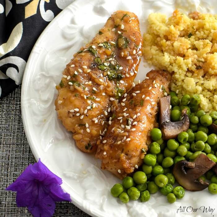 Close up of two honey glazed chicken tenders along with golden quinoa and peas with mushrooms all on a white edge embossed plate. A purple blossom is on the lower left hand edge of the plate and a black napkin with white fleur de lis on the upper left side. 