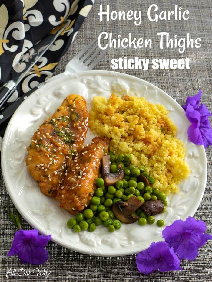 Two pieces of honey garlic chicken thighs on a white plate with green peas and mushrooms, and golden quinoa. The white fruit embossed plate is surrounded by 3 purple blossoms, on a brown woven placemat with a knife and fork on the side and a black, white, and gold napkin. 