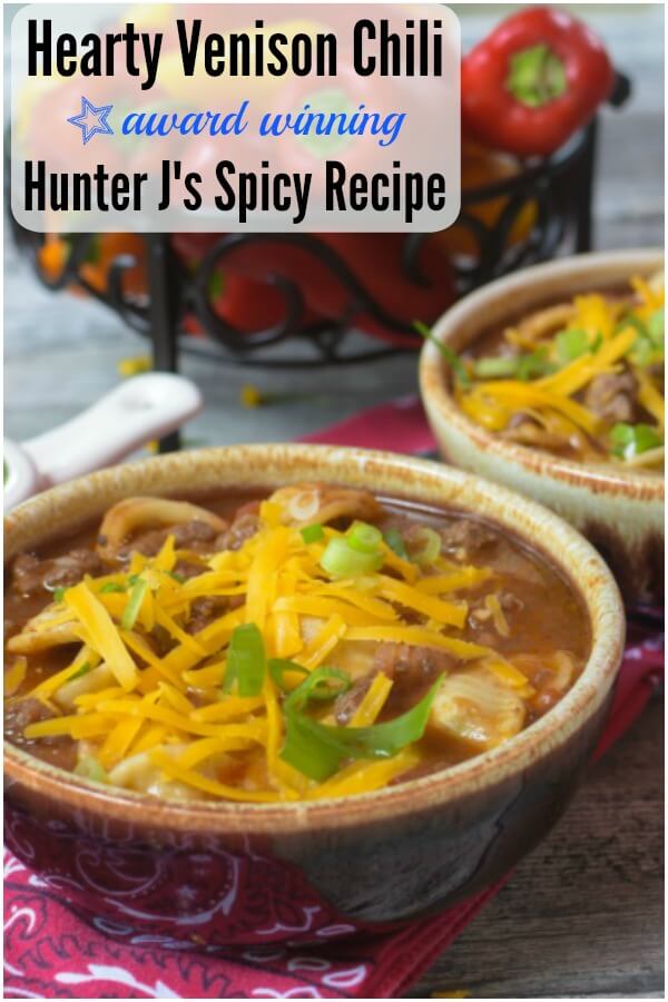 Two bowls of hearty venison chili in brown glazed bowls atop of red bandanas. Shredded yellow cheese and green onion circles top the chili. Red and yellow miniature peppers in a wrought iron container in background. 