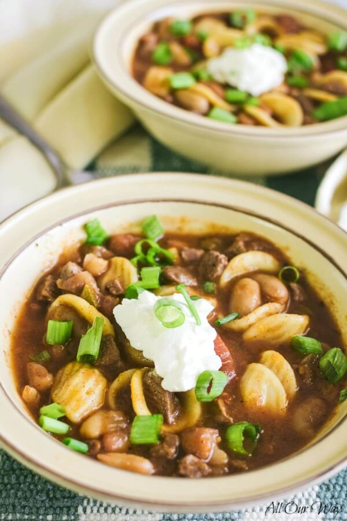 A closeup of chili con carne with beans and round pasta. There is a dollop of sour cream in the middle of the bowl and a sprinkle of chopped green onions on top. Part of another bowl of chili is behind the first bowl. A soup spoon is off to the left side.