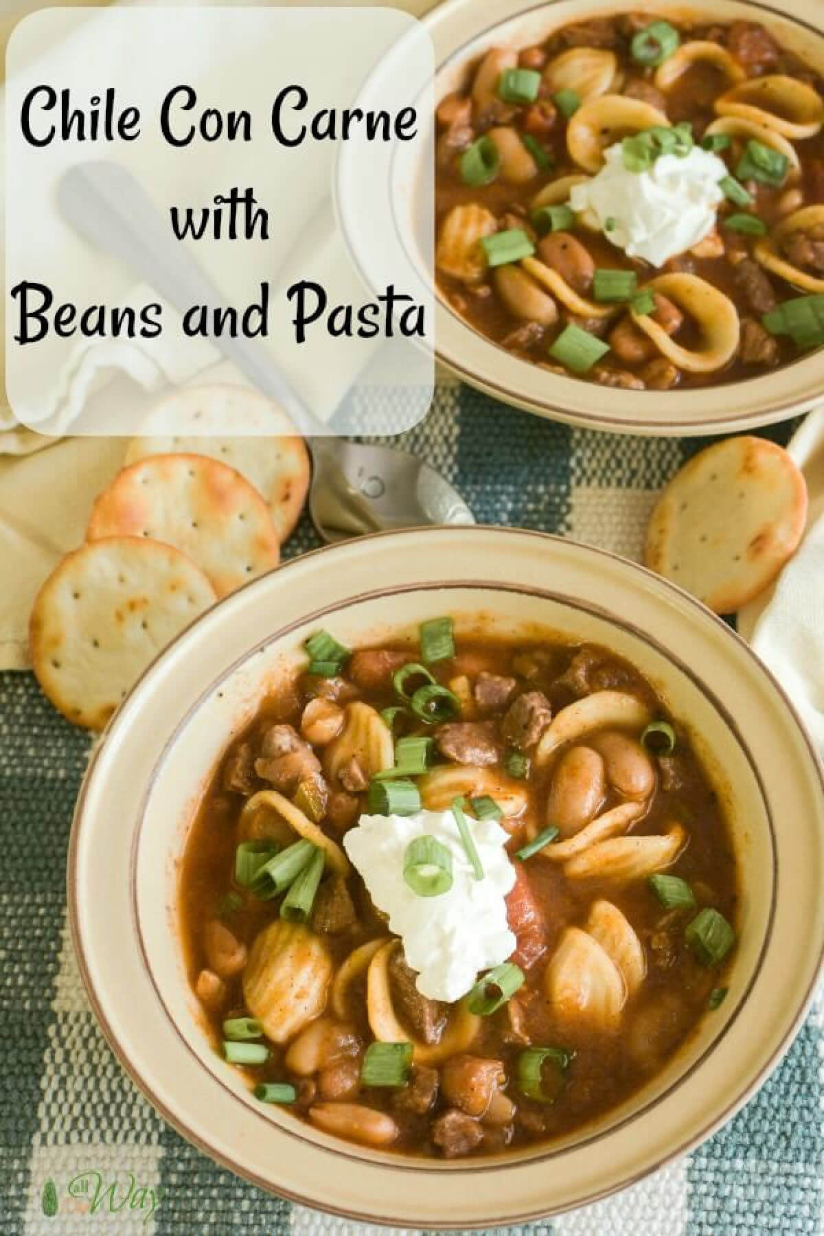 Two bowls of chili con carne with round pasta and beans topped with sour cream and a sprinkle of green onion rings. Round crackers are around bowls all sitting on a green and white plaid placemat.