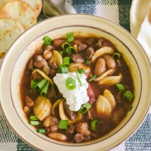 Overhead view of two yellow bowls of chili con carne with dollops of sour cream in the center of the soup and a sprinkle of green onions over the top. There are round pita crackers surrounding the bowl and a silver soup spoon is on the left side. The bowls are on green and white plaid placemat.