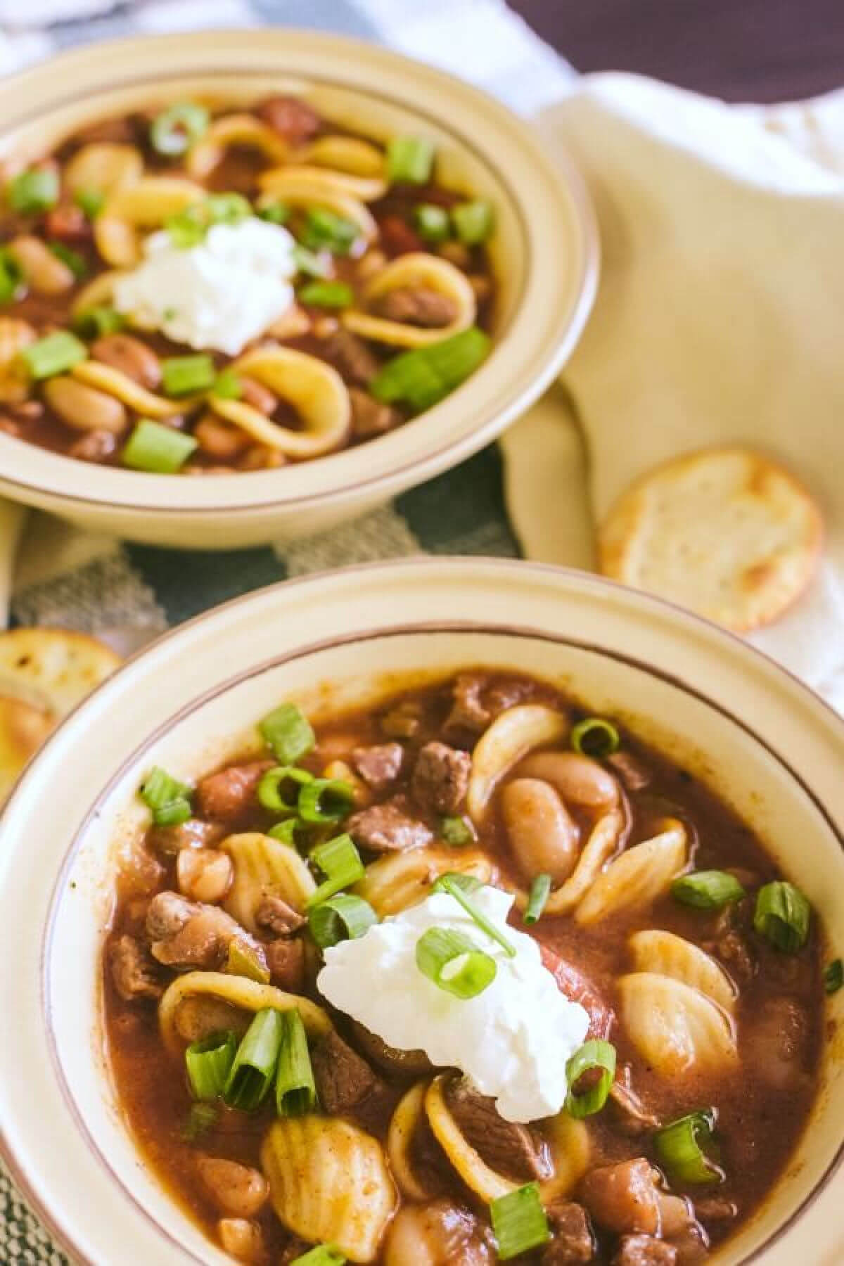 An overhead closeup of two yellow bowls of chili_con_carne with dollops of sour cream in the middle of each bowl and sprinkling of green onions over the top. The bowls are on white and green plain placemat and three pita crackers next to the bowls.