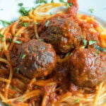 Three baked venison meatballs with spaghetti and red sauce on a white porcelain plate all on top of a green and white checked placemat. A fork rests on the left hand side of the plate.