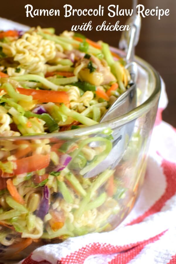 A large clear glass bowl filled with broccoli coleslaw with bits of cooked chicken and matchstick pieces of red bell pepper. The bowl rests on a red and white tea towel and silver salad serving fork in the bowl.