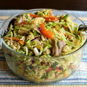 A clear glass bowl filled with ramen broccoli slaw on top of a blue, yellow, white striped tea towel.