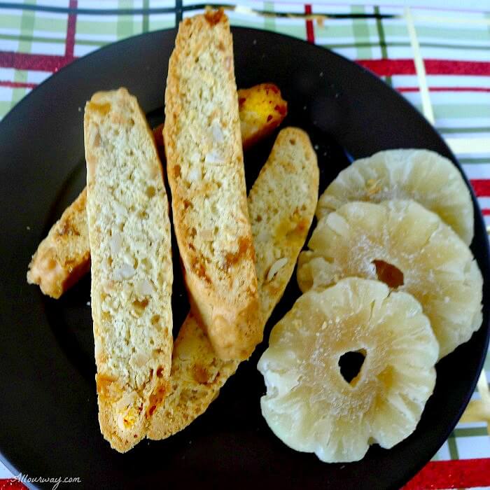 Piña Colada Biscotti stacked on a brown plate with slices of dried pineapple all on green and red Christmas wrapping paper. 