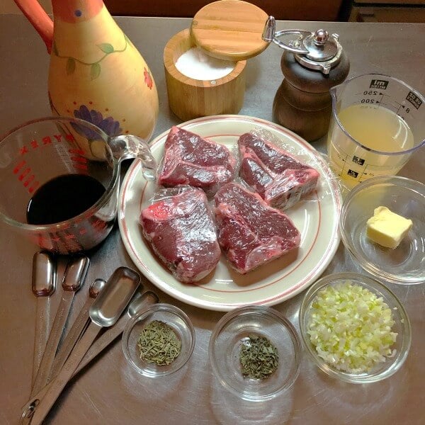 Ingredients for the Balsamic Glazed lamb chops: 4 lamb chops on white plate, balsamic vinegar in cup, olive oil in yellow flowered bottle, sea salt in wooden container, measuring spoons, chicken broth in cup, pat of butter in glass dish, chopped shallots in glass prep bowl, dried rosemary in glass dish, dried time in glass dish. 