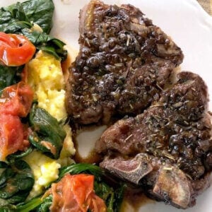 Two browned lamb chops on a white plate with yellow polenta covered with sauteed red grape tomatoes and green baby spinach.