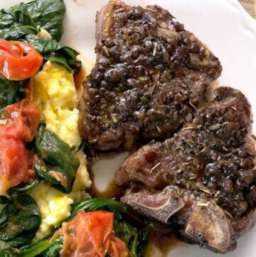 Two browned lamb chops on a white plate with yellow polenta covered with sauteed red grape tomatoes and green baby spinach.