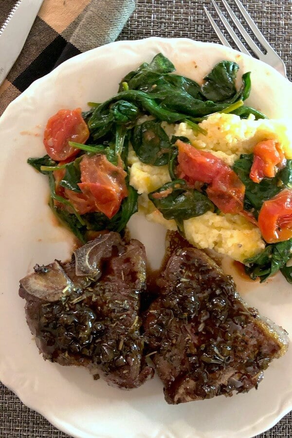 Two balsamic glazed lamb chops on a white plate with yellow polenta and sautéed baby spinach and grape tomatoes on top. 