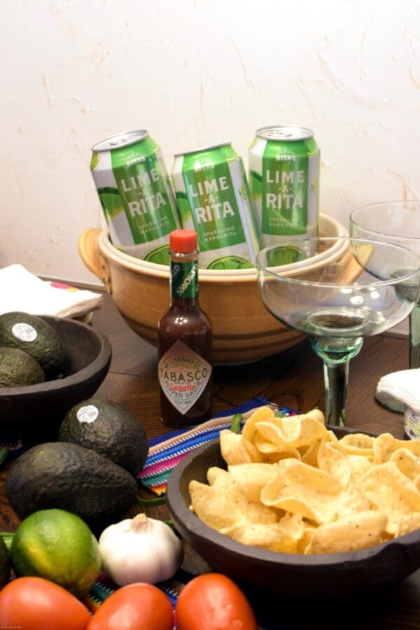 A wooden party table with avocados, tortilla scooper chips, green and white cans chilling in brown pottery pot, a green margarita glass and a bottle of TABASCO® Chipotle Sauce.