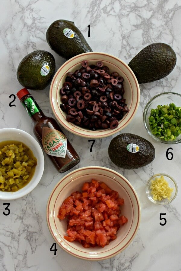 Ingredients for Italian Avocado Salsa on marble, six avocados, TABASCO® Chipotle Sauce, green chilies in white bowl, diced tomatoes in bowl, minced garlic in glass prep dish, chopped green onions in glass, sliced black Kalamata olive in white bowl.