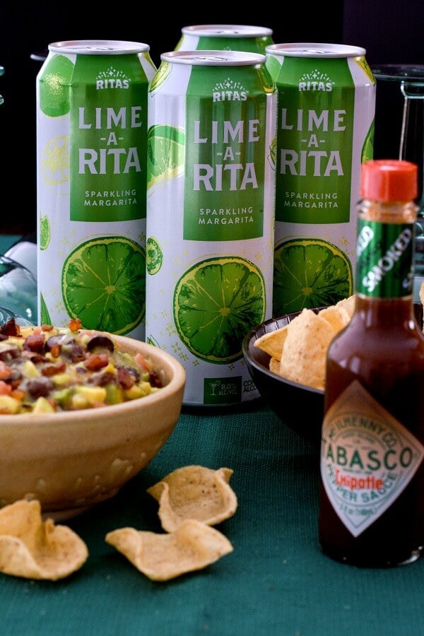 Tan pottery bowl holds Italian avocado salsa, 3 green and white aluminum cans of Lime-A-Rita , bottle of TABASCO® Chipotle Sauce and bowl of tortilla chips on a green tablecloth. 