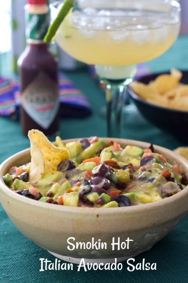 Close up of a tan pottery bowl tilled with Italian avocado salsa a mixture of avocados, diced tomatoes, black olives, and a tortilla chip stuck in the salsa, with a bottle of TABASCO® Chipotle Sauce in the background and a green margarita glass filled with Lime-A-Rita drink. 