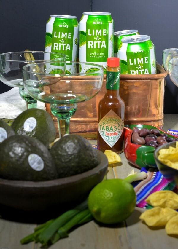 Avocados in wooden bowl with lime, TABASCO®Sauce, green and white aluminum Lime-A-Rita cans in copper tub with green margarita glasses and tortilla chips on table. 