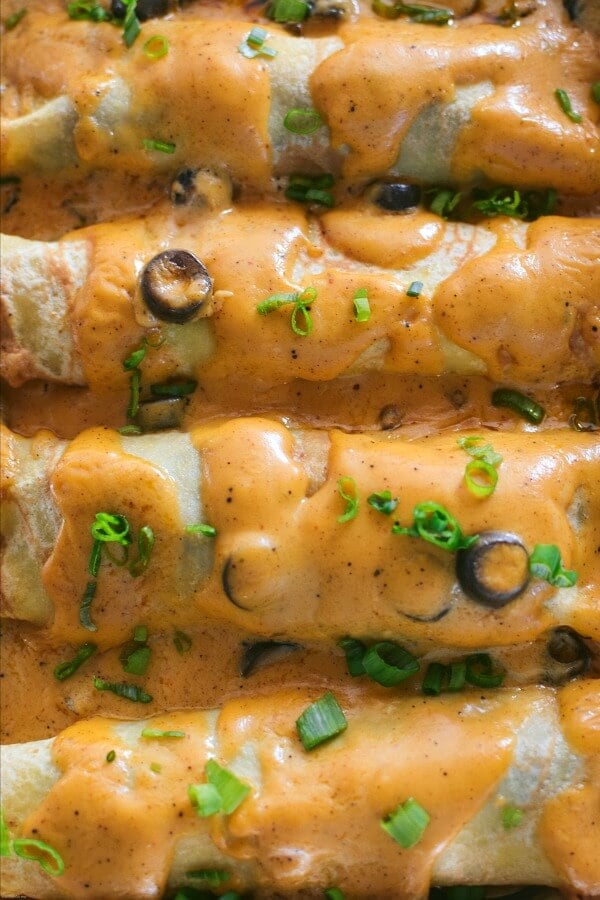 Close up cheese sauce covered beef enchiladas lined up in a aluminum casserole pan. 