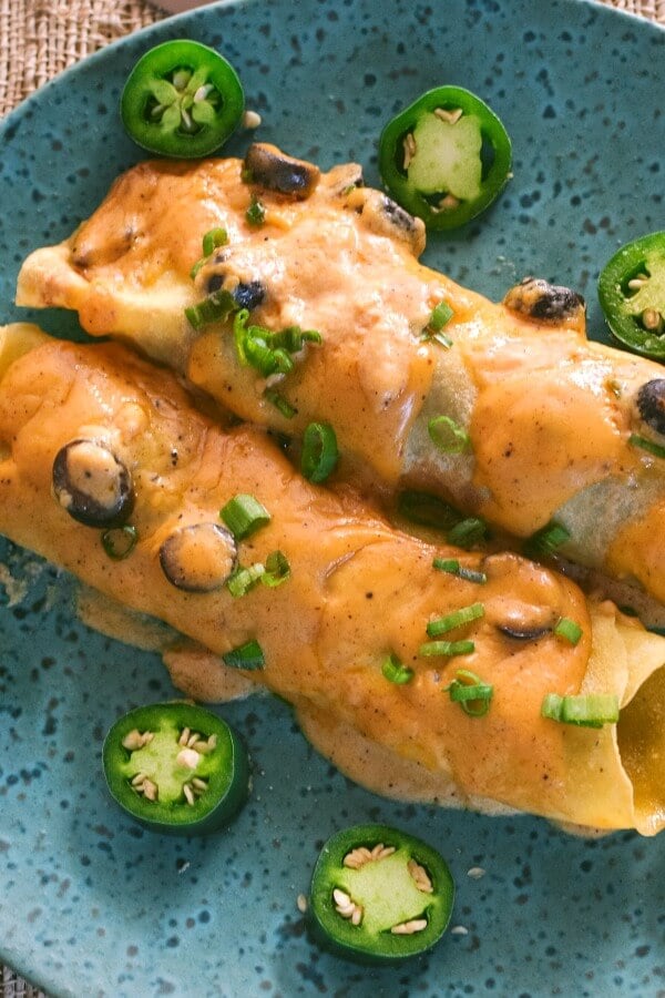 Two Cheese covered beef enchiladas on a teal speckled pottery plate with slices of green jalapeños surrounding the enchiladas. 