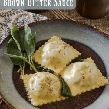 Three lamb ravioli on a blue and brown round plate with a sprig of sage on the side. A multi-color napkin is under the plate.