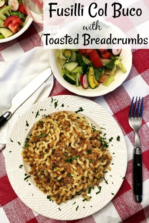 Overhead view of a white bowl filled with fusilli pasta sprinkled with toasted breadcrumbs and a2 small tossed salads next to the bowl and a glass of white wine and black handled dinnerward. 