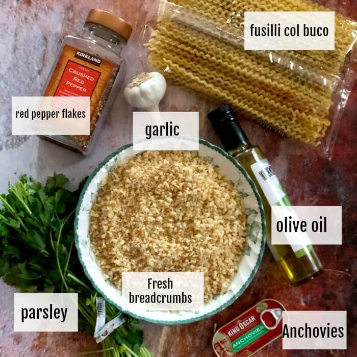 Overhead view of the ingredients for Fusilli Col Buco With Toasted breadcrumbs including, hot pepper flakes, parsley, garlic, fusilli pasta, olive oil, and a tin of anchovies.