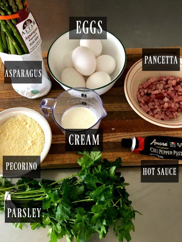 An overhead shot of a frittata ingredients, eggs, prociutto, asparagus, pecorino-romano cheese, cream, hot sauce paste, parsley. 