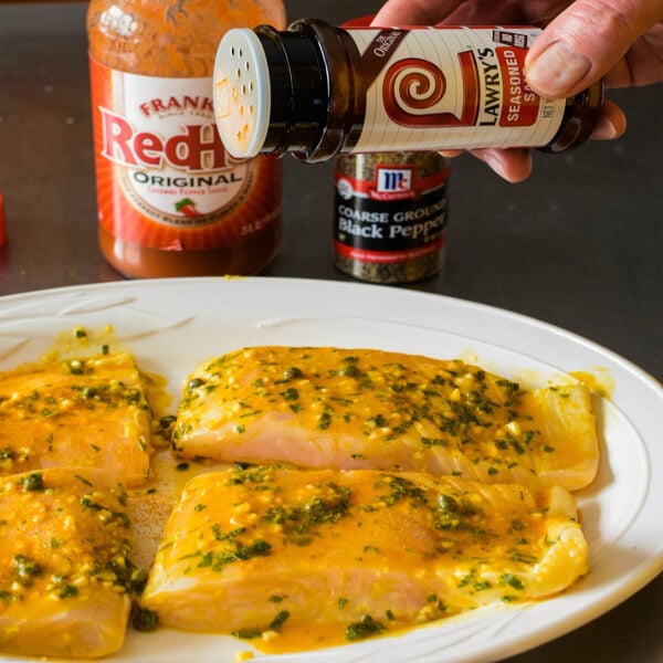 Four halibut fillets on a platter being seasoned with Lawry's®Seasoned Salt and hot sauce and pepper in the background. 