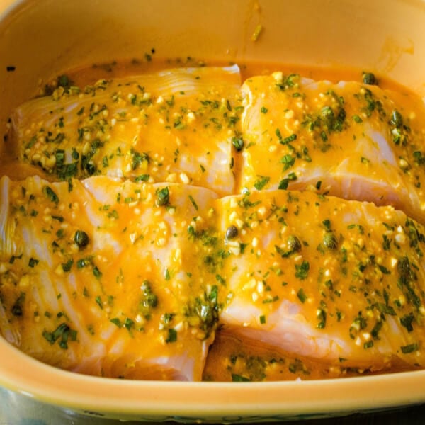 Four halibut fillets marinating in a herb citrus marinade in a gold glass casserole. 